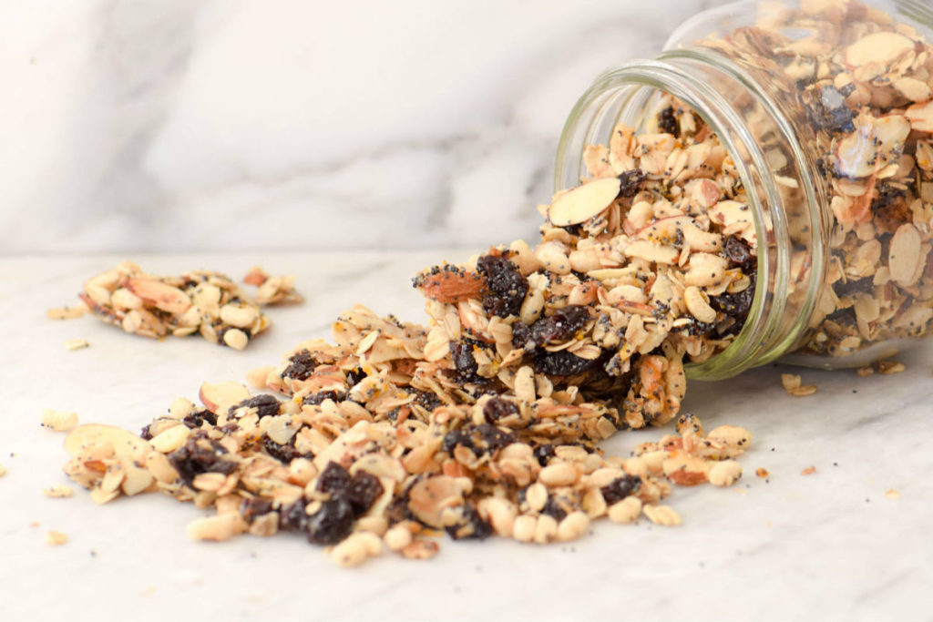 Granola casera con avena, frutos secos y pasas saliendo de un frasco de vidrio sobre superficie blanca.