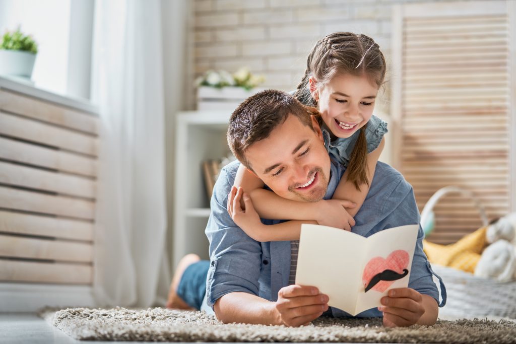 Padre sonriendo mientras lee una tarjeta hecha a mano que le regaló su hija, quien lo abraza felizmente por el Día del Padre.