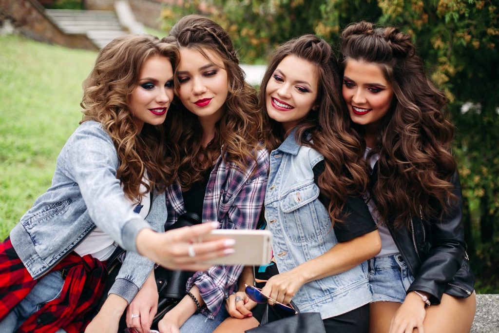 Cuatro amigas sonrientes con maquillaje de día natural tomándose un selfie al aire libre