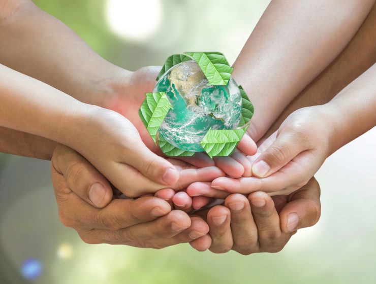 Varias manos sostienen una figura del planeta Tierra rodeada de hojas verdes, representando el cuidado del medio ambiente.