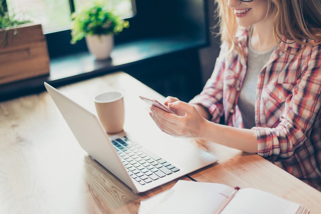 mujer sonriente usando smartphone junto a laptop y taza de café en escritorio de madera