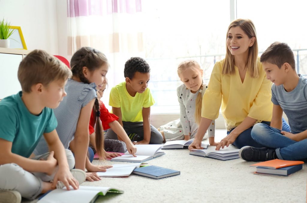 Maestra joven sentada en el piso con sus alumnos leyendo libros